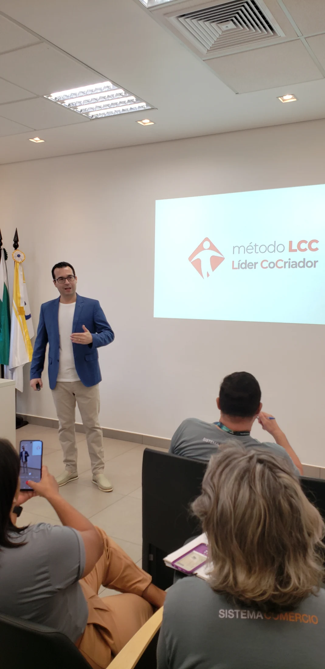 Gustavo Isaias apresenta diante de uma projeção, com participantes em primeiro plano